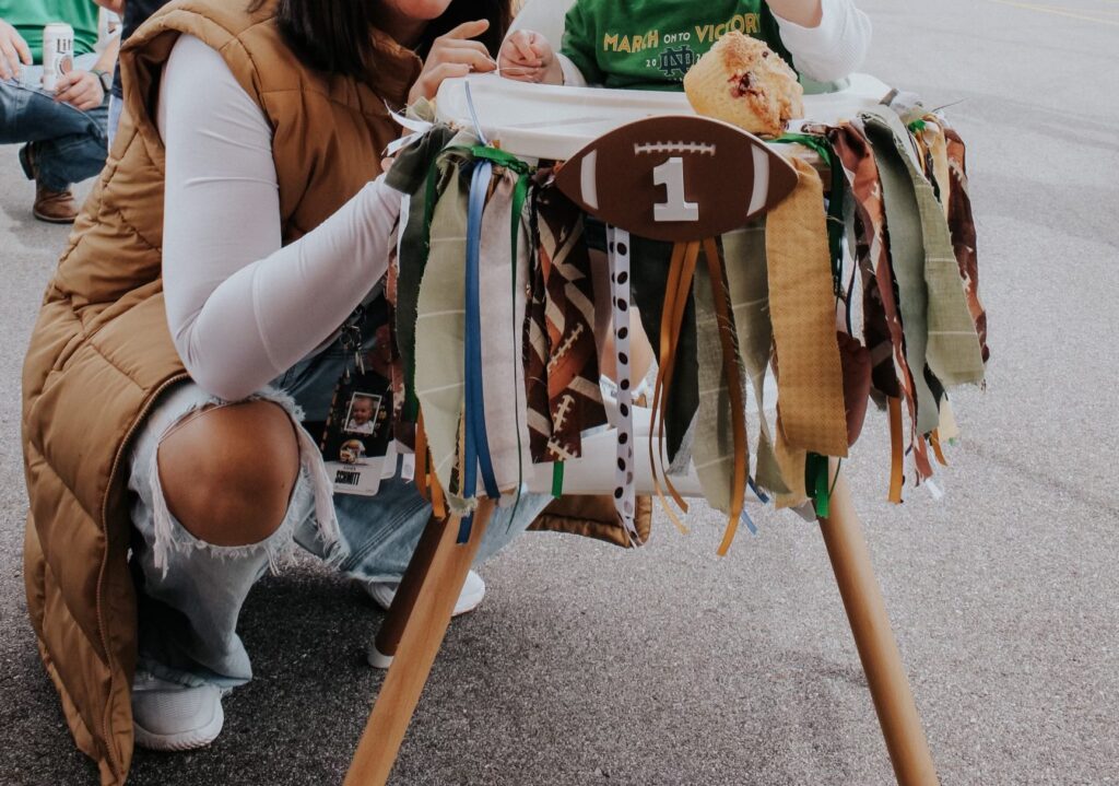 High chair garland that is football themed with a football in the middle with a 1 on it and yellow and green ribbon, hanging off of a highchair.