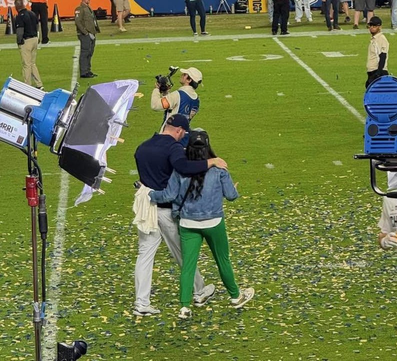 husband and wife walking away from the camera on a football field hugging each other.