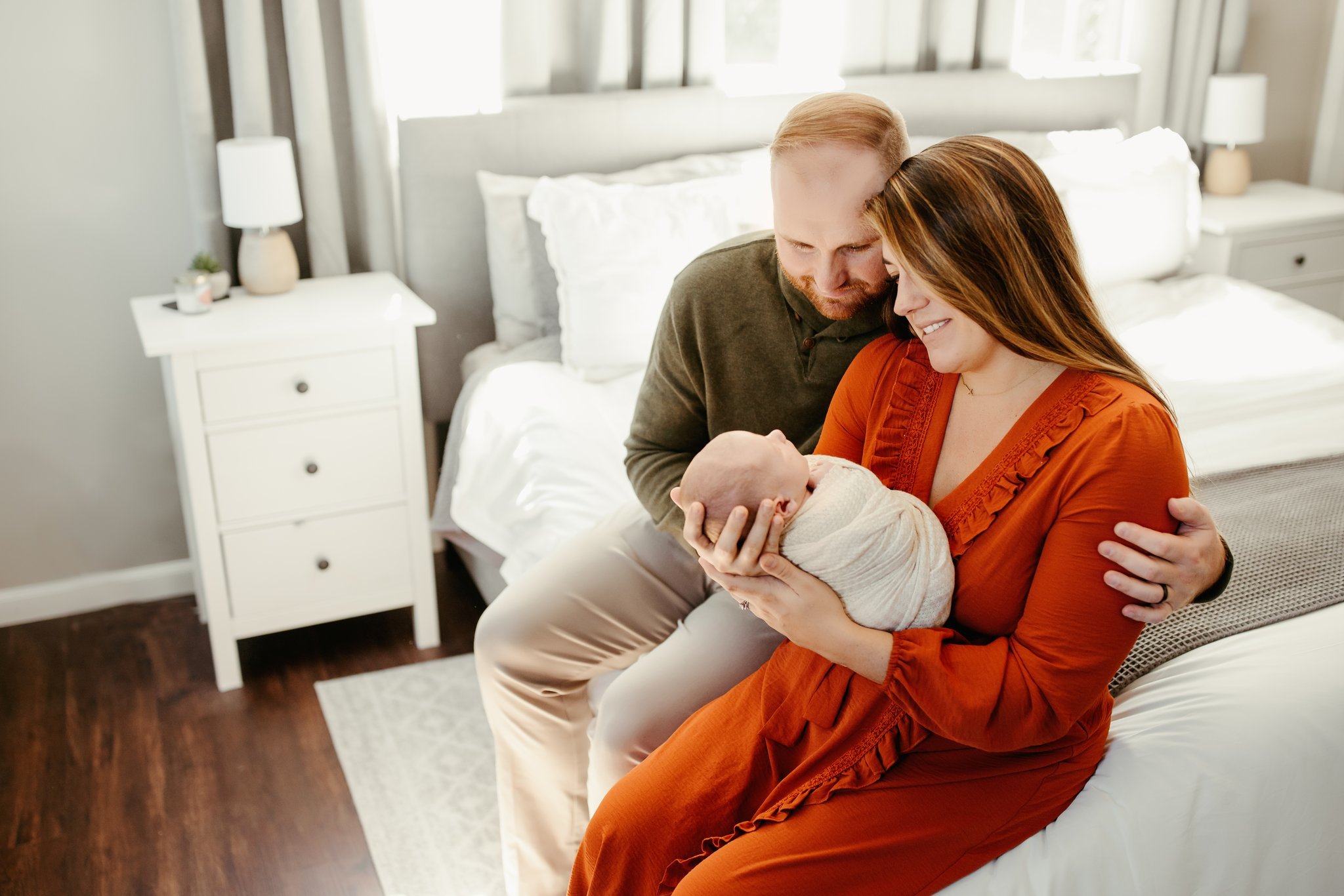 A mom and dad holding their newborn in their cozy home décor, swaddled in blankets.