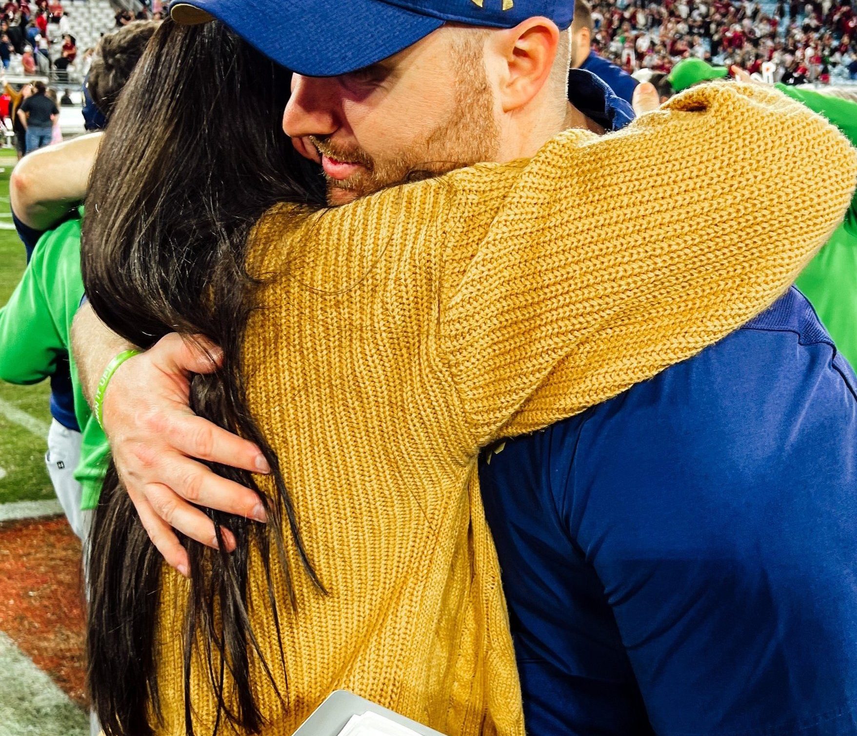 Picture of a husband and wife hugging at a football game on the field with fans in the background.