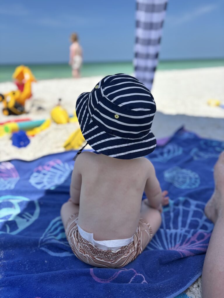 A baby with a bucket hat sitting on a beach with the baby's back towards the camera.