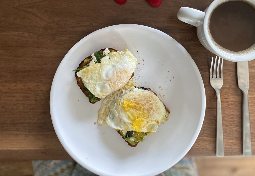Top-down view of avocado toast on with egg on top on a plate, with a cup of coffee next to silveware on a table.
