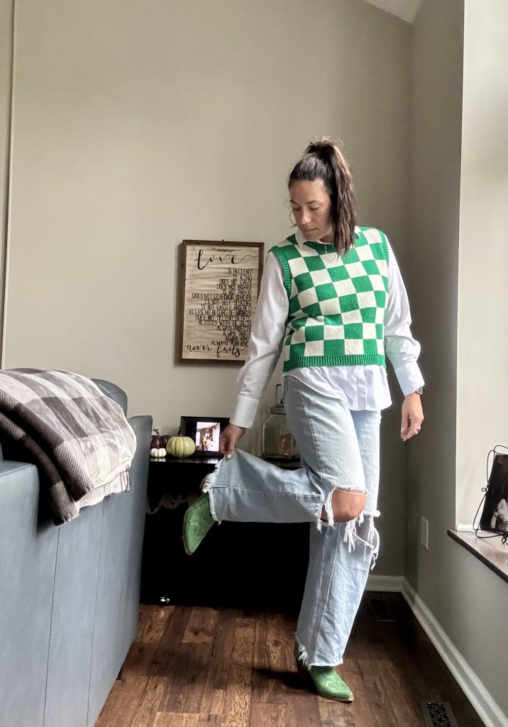 Woman standing in jeans, a button-down, with a checkered Kelly green sweater vest, and green cowboy boots.
