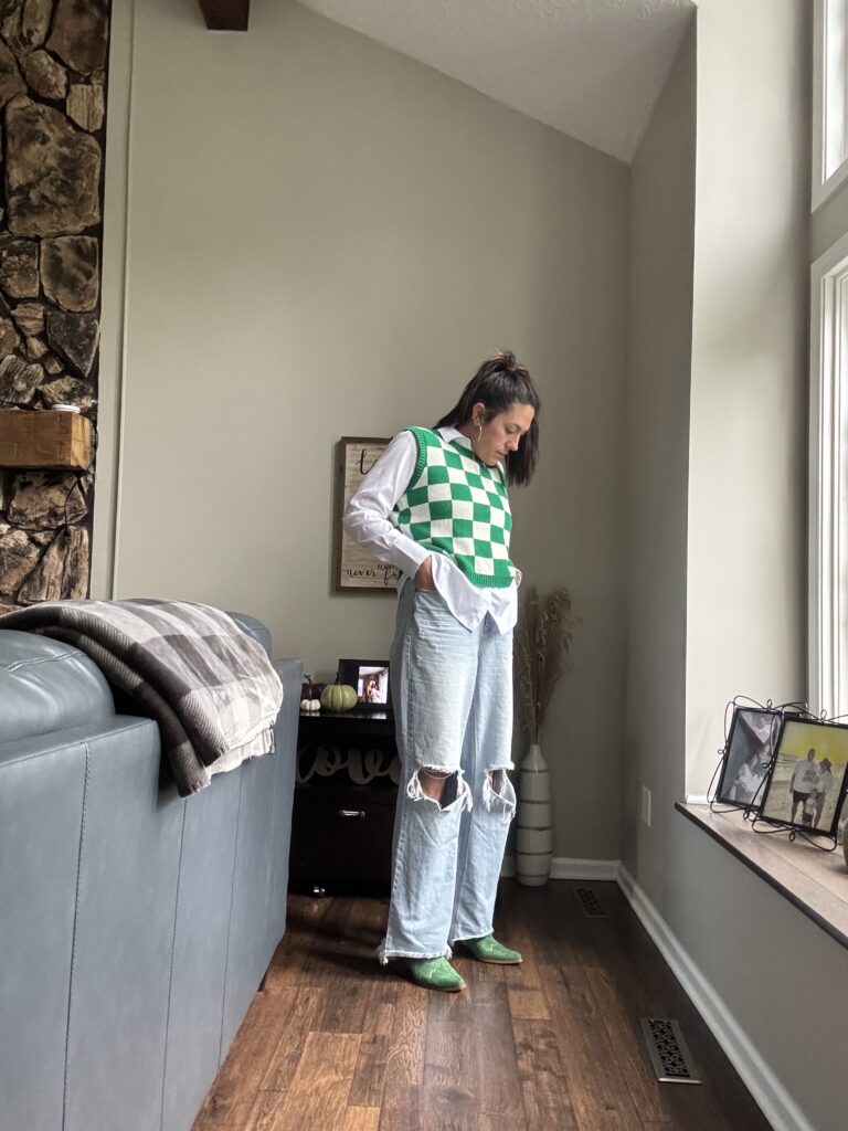 Woman standing next to a window in a kelly green sweater vest, jeans, and green booties.