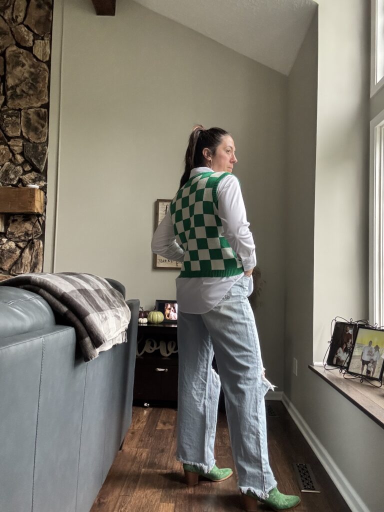 Woman standing in jeans, green cowboy boots, button down blouse and a green checkered sweater vest.