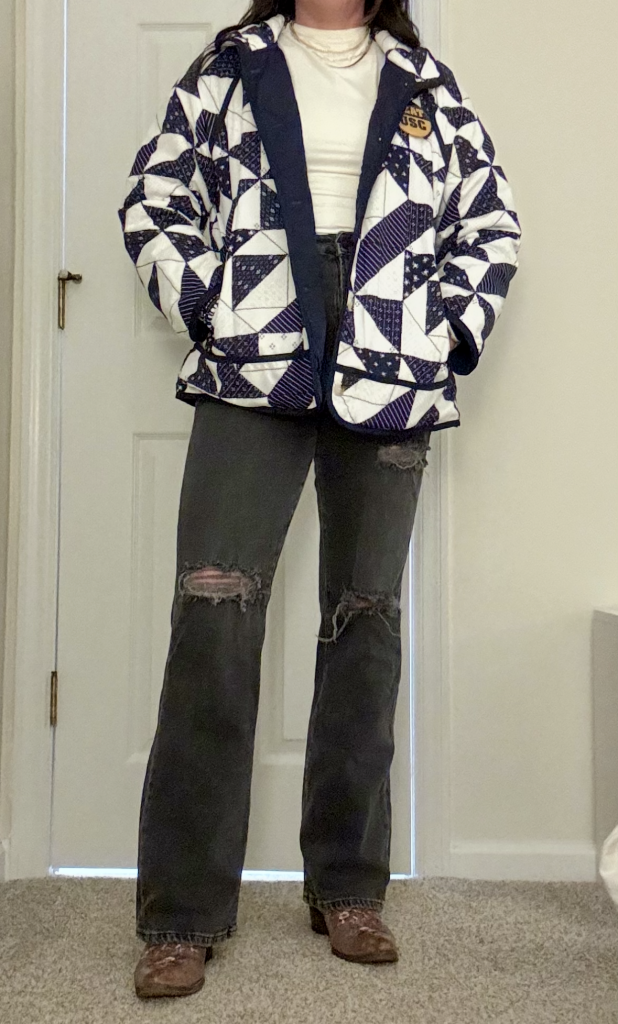 Profile of a woman standing in black jeans, boots, and navy quilted jacket, with layered necklaces.