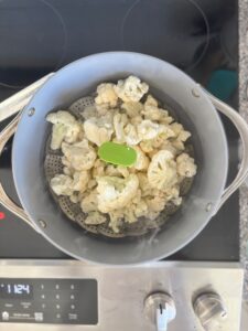 Top-down view of chopped up cauliflower in a steamer in a pot of water on a stove.
