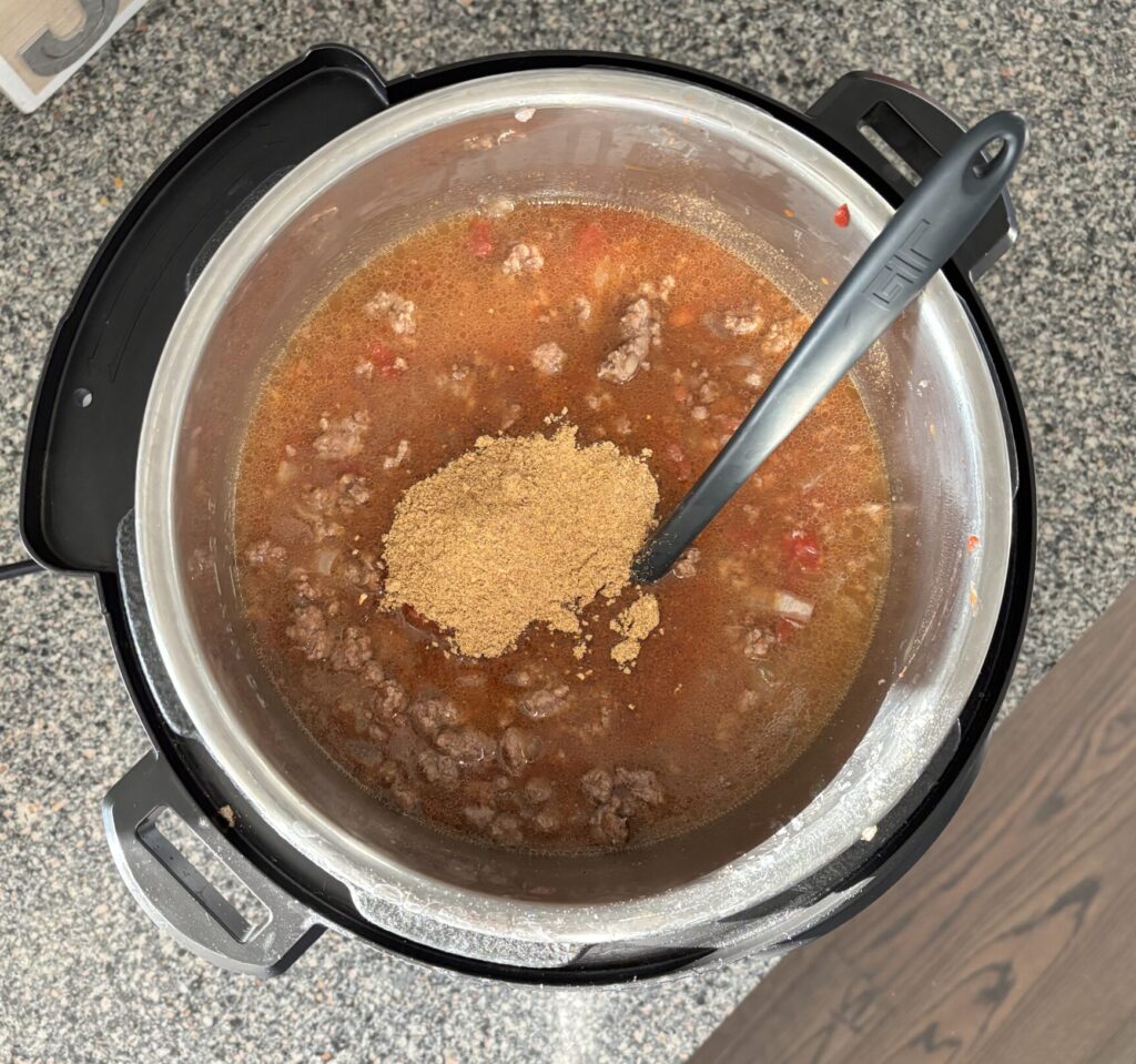 Top-down view of low-carb chili ingredients in an Instant Pot on the soup setting, including tomato paste, diced chili-ready tomatoes, mild chili powder seasoning packet, Chili beans, and bone broth.
