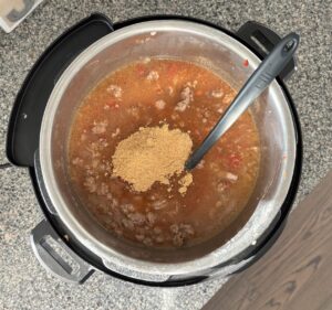 Top-down view of low-carb chili ingredients in an Instant Pot on the soup setting, including tomato paste, diced chili-ready tomatoes, mild chili powder seasoning packet, Chili beans, and bone broth.