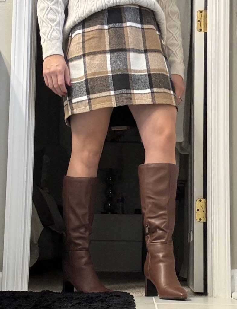 Waist down view of plaid skirt and chocolate brown boots dressed with a cream knit sweater that is tucked in.