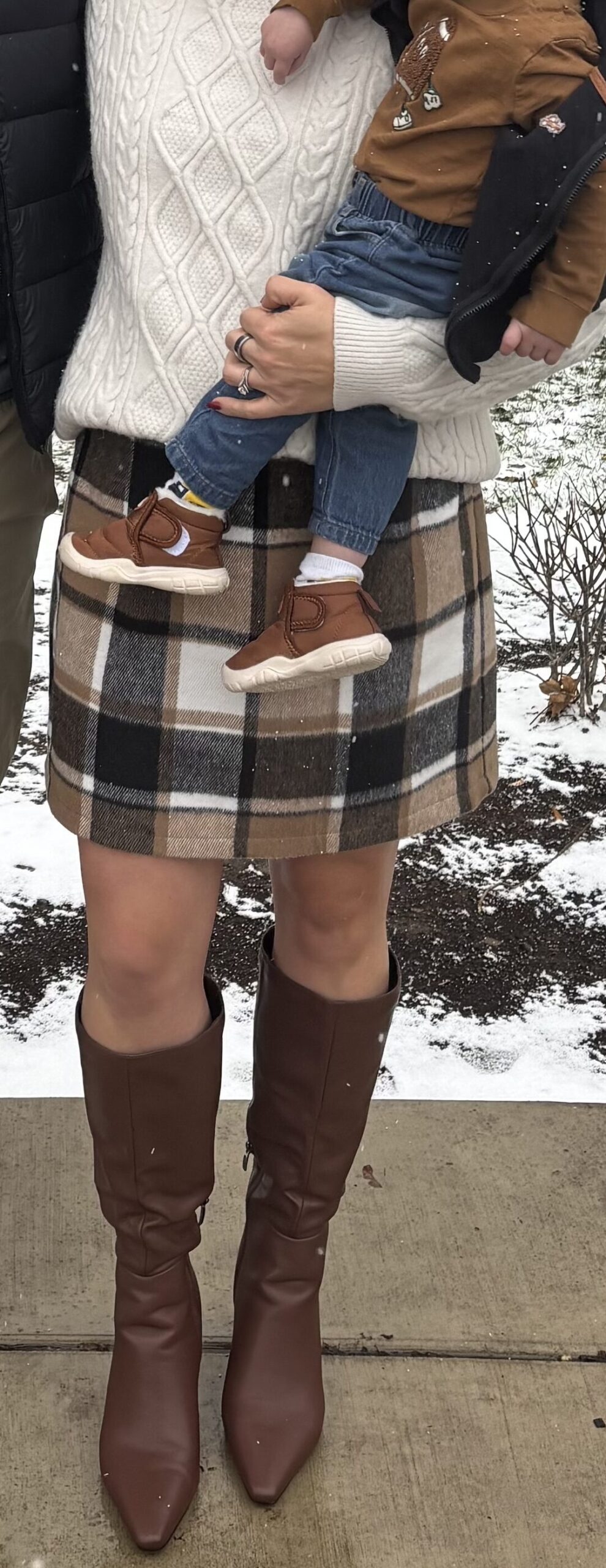 Woman wearing a winter plaid skirt with chocolate brown boots while holding a child.